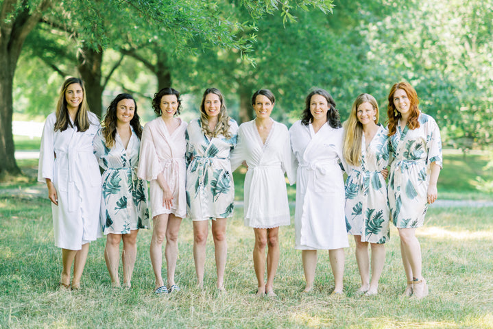 Eight women stand in a row outdoors on grass under green trees, smiling. They wear light-colored robes, with some floral patterns, and appear to be part of a wedding party or getting ready for an event.