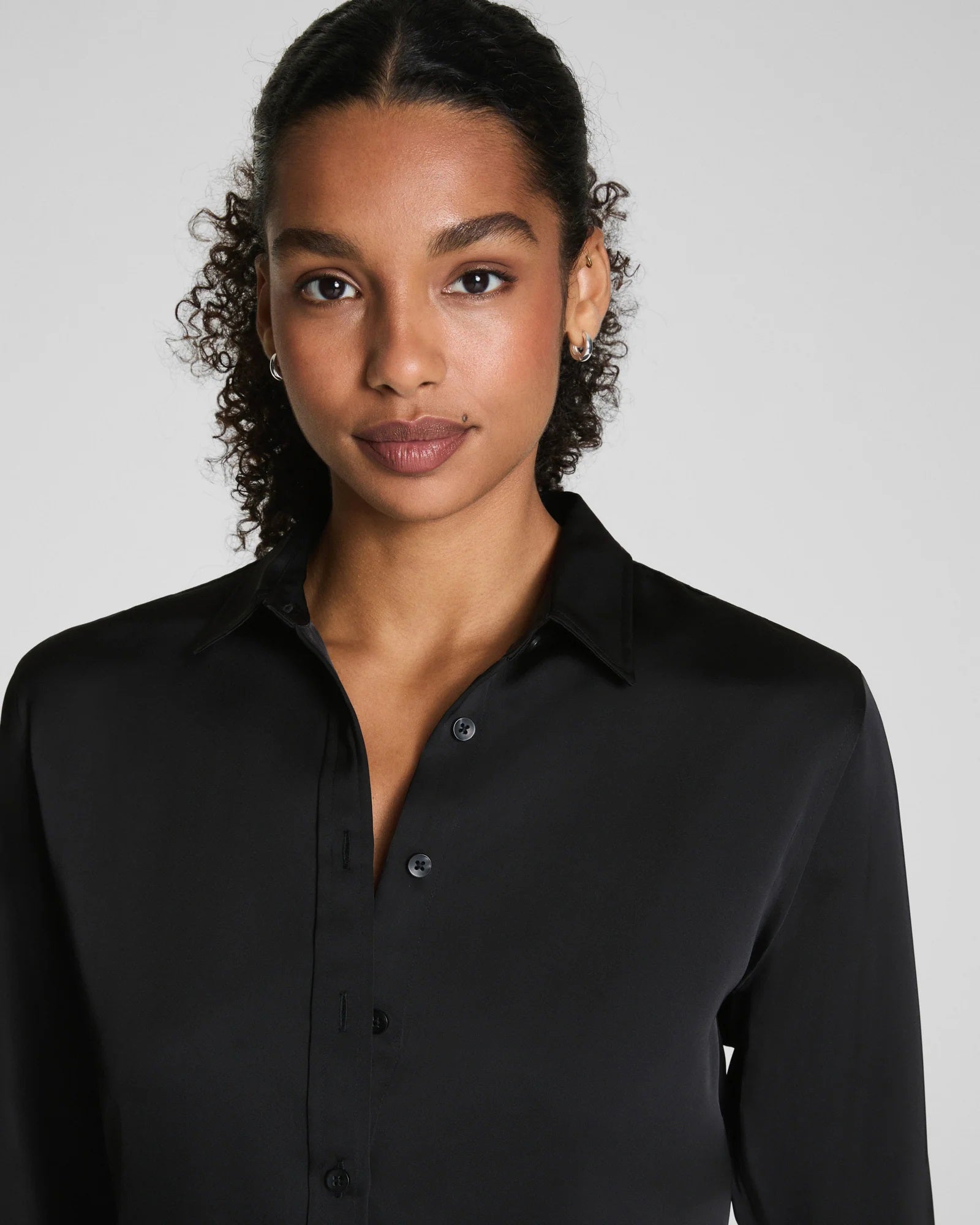 A woman with curly hair pulled back wears the SPANX Silky Smooth Button Down Shirt in black, paired with black pants. She stands facing away, looking over her shoulder against a plain light gray background.