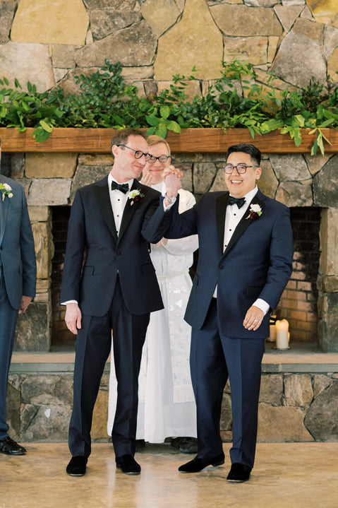 Two grooms in tuxedos hold hands and smile joyfully at each other during their wedding ceremony, standing in front of a stone fireplace with greenery, as an officiant stands behind them.