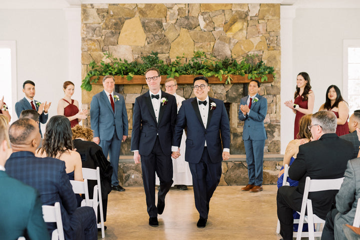 Two grooms in tuxedos walk hand-in-hand down the aisle, smiling, after their wedding ceremony. Guests seated on either side clap and celebrate. The setting features a stone fireplace with greenery.