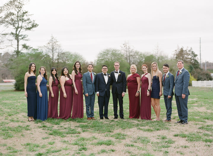 A wedding party poses outdoors on grass, with six women in burgundy and navy dresses and five men in suits, standing side by side. Trees and a cloudy sky form the background.