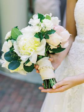 A bride in a white lace dress holds a bouquet of white and pale pink flowers, wrapped with a cream ribbon and adorned with pearl accents.