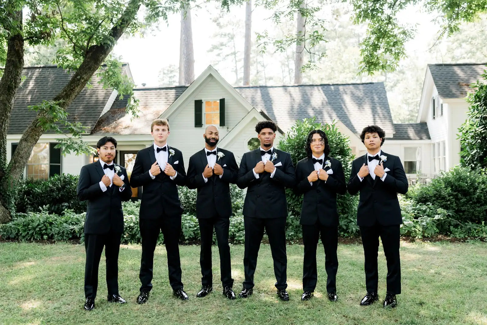 Six men in black tuxedos stand in a row on a grassy lawn, smiling and holding their jacket lapels, with a white house and trees in the background.