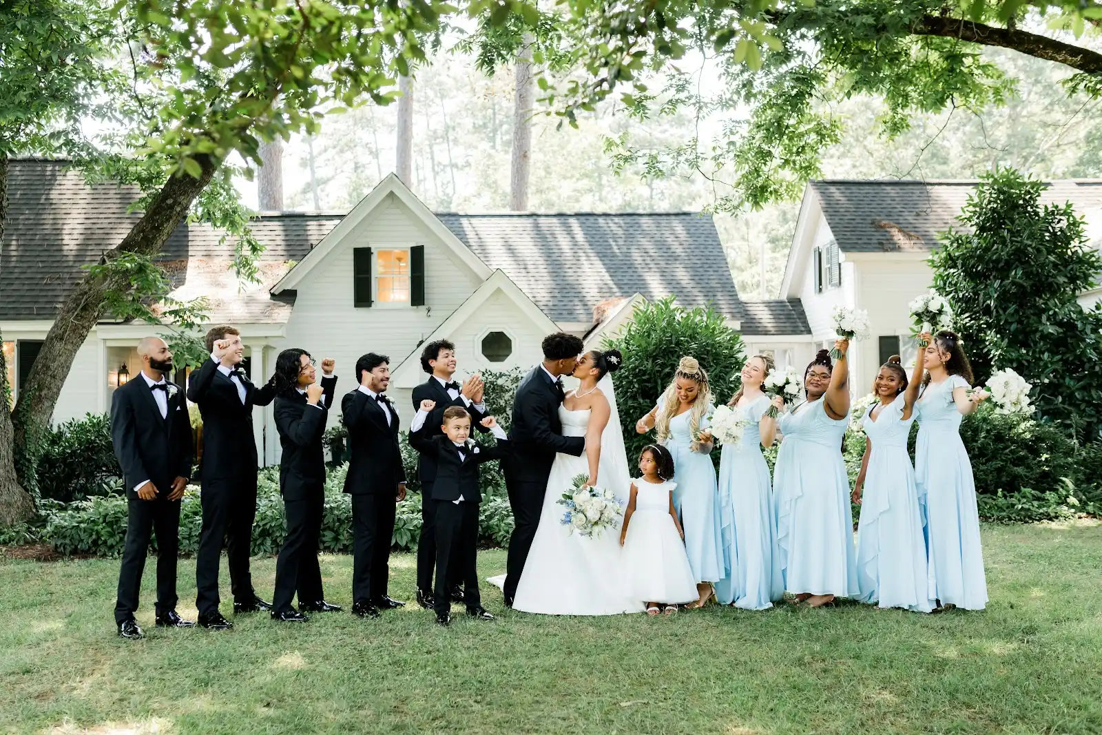 A wedding party poses outdoors in front of white houses. The bride and groom stand in the center, surrounded by bridesmaids in light blue dresses and groomsmen in black suits, all celebrating and smiling.