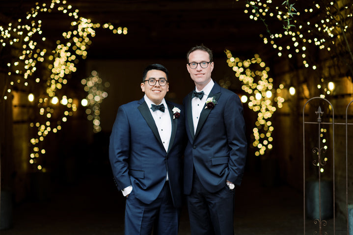 Two men in tuxedos stand side by side, smiling, in a warmly lit venue with string lights and blurred bokeh in the background, creating a festive and elegant atmosphere.