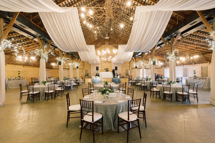 A rustic barn venue decorated for an event, featuring round tables with white tablecloths, wooden chairs, draped white fabric on the ceiling, and hanging string lights with greenery accents.