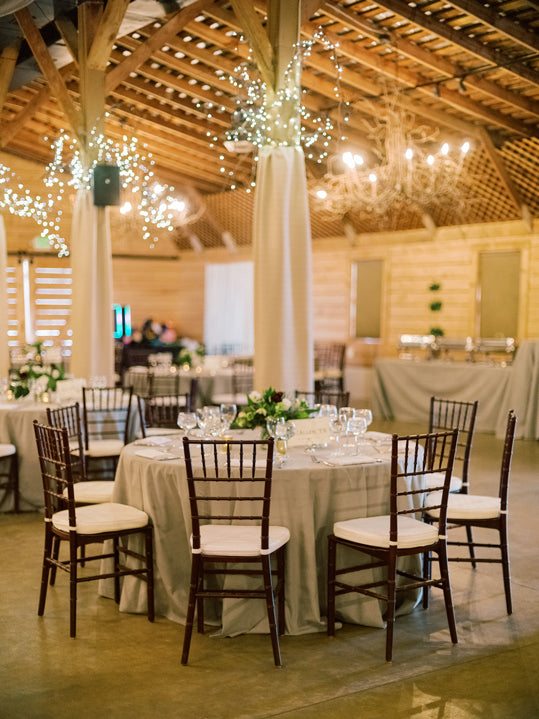 A round table set for six with a white tablecloth, floral centerpiece, and glassware in a rustic venue with wooden beams, string lights, and a chandelier overhead. Empty wooden chairs surround the table.