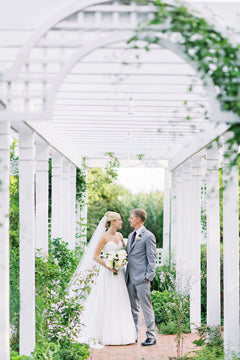 A bride and groom stand under a white garden pergola, surrounded by greenery. The bride wears a white dress and veil, holding a bouquet, while the groom wears a light gray suit. They look at each other and smile.