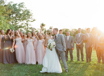 A bride and groom kiss outdoors as their smiling wedding party, dressed in pastel dresses and gray suits, celebrates around them in a sunlit grassy field.