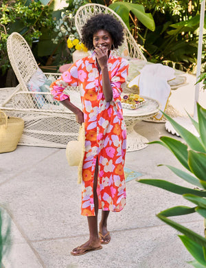 A person with curly hair stands outdoors, wearing the FRANK & EILEEN Rory Maxi Shirtdress in Classic Italian Linen—Orange and Pink Floral—embodying California chic amid lush greenery and white outdoor furniture, holding a woven straw bag.