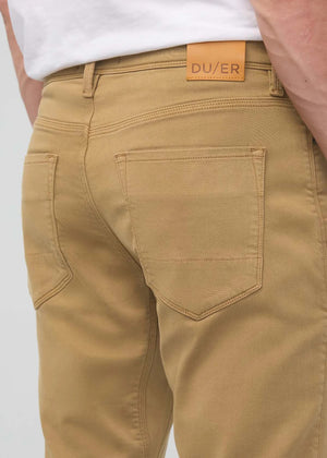 A man with short dark hair and a trimmed beard stands against a white background, wearing a white t-shirt, white sneakers with black details, and DUER's DU/ER NO SWEAT RELAXED TAPER PANT in an athletic fit. One hand is in his pocket.
