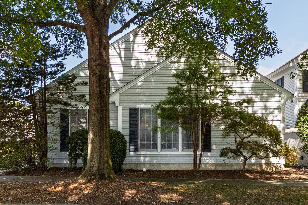 A white house with large front windows, black shutters, and several trees and bushes in the yard casting shadows on the facade.