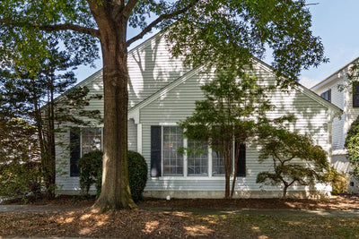A white house with large front windows, black shutters, and several trees and bushes in the yard casting shadows on the facade.