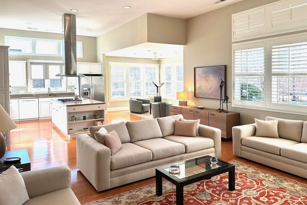 A bright, modern living room with light-colored sofas, a glass coffee table on a red patterned rug, large windows with white shutters, and an open kitchen in the background.