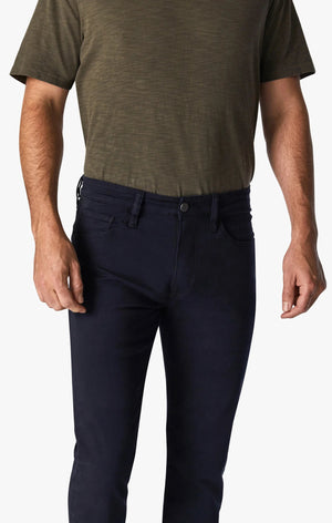 A man with short hair and a beard stands facing forward, wearing an olive green T-shirt, 34 HERITAGE - COURAGE STRAIGHT LEG PANT IN NAVY TWILL, and brown shoes against a plain white background.
