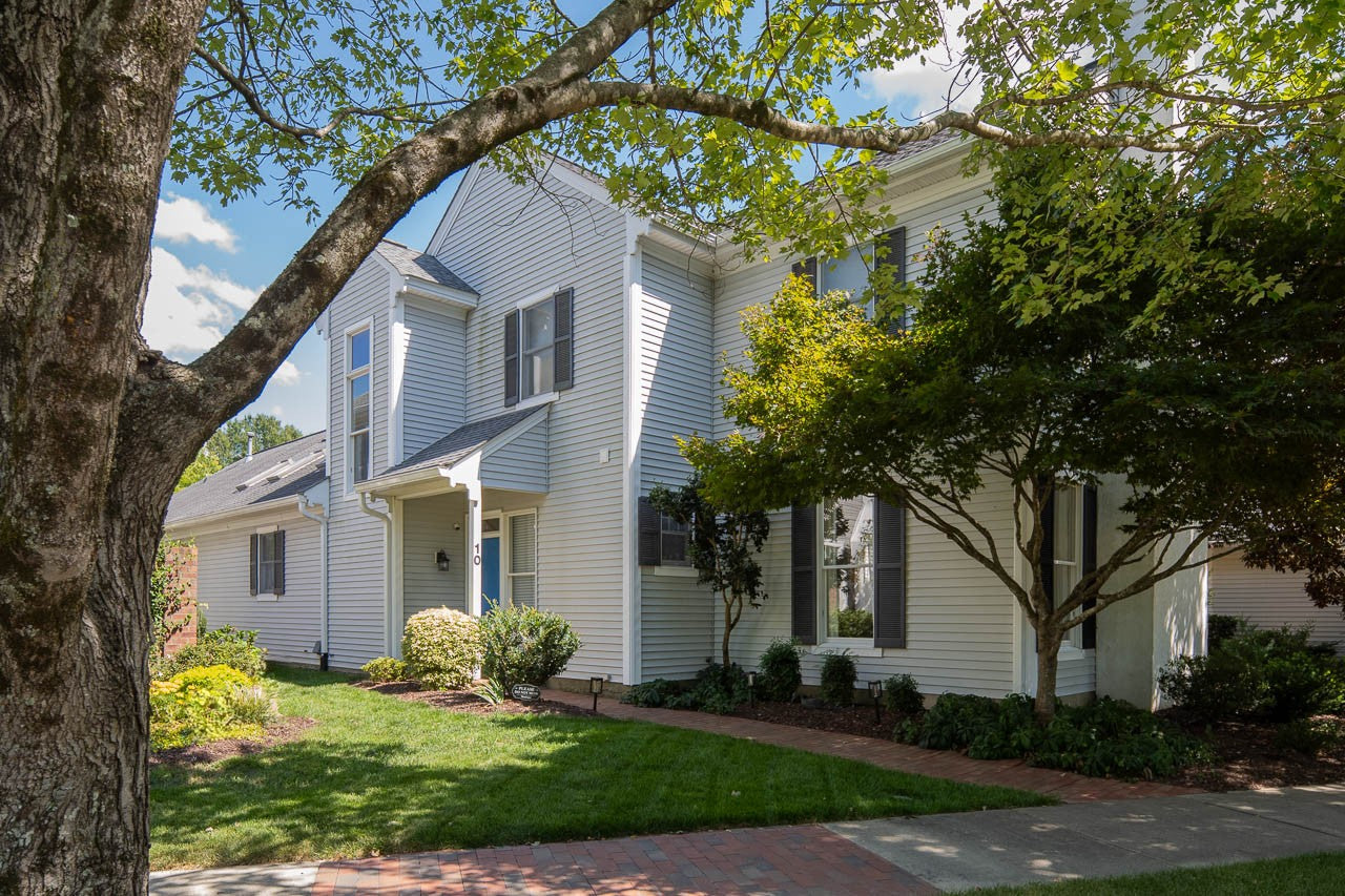 A two-story white house with dark shutters and a front porch, surrounded by green grass, shrubs, trees, and a brick sidewalk under a blue sky.