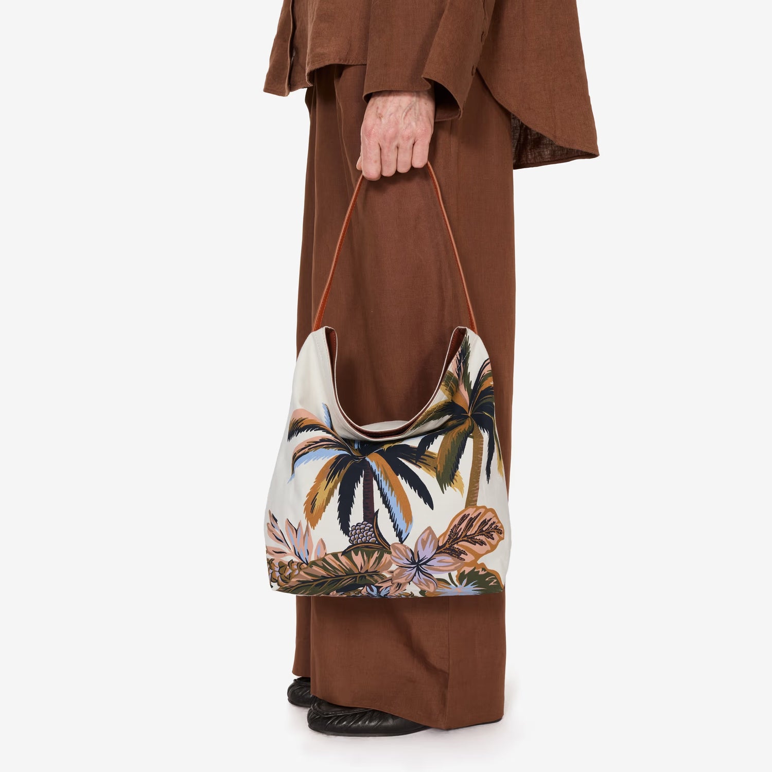 A person dressed in loose brown clothing holds a large, white tote bag with a tropical palm tree and foliage print. The bag hangs from their hand, and only their lower body is visible.