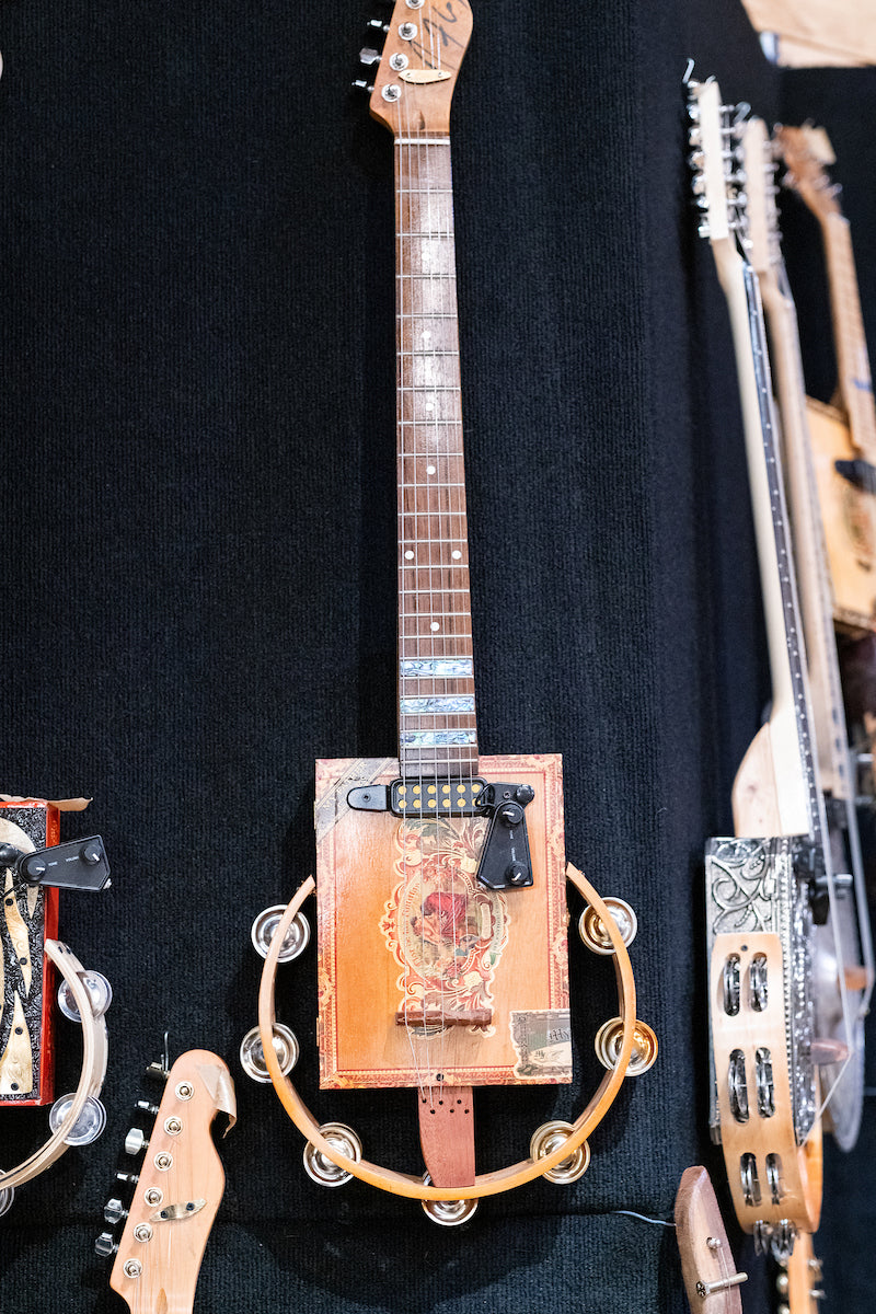 A handmade guitar with a wooden cigar box body, metal string hardware, and a tambourine attached around the base, displayed vertically against a dark background among other unique string instruments.