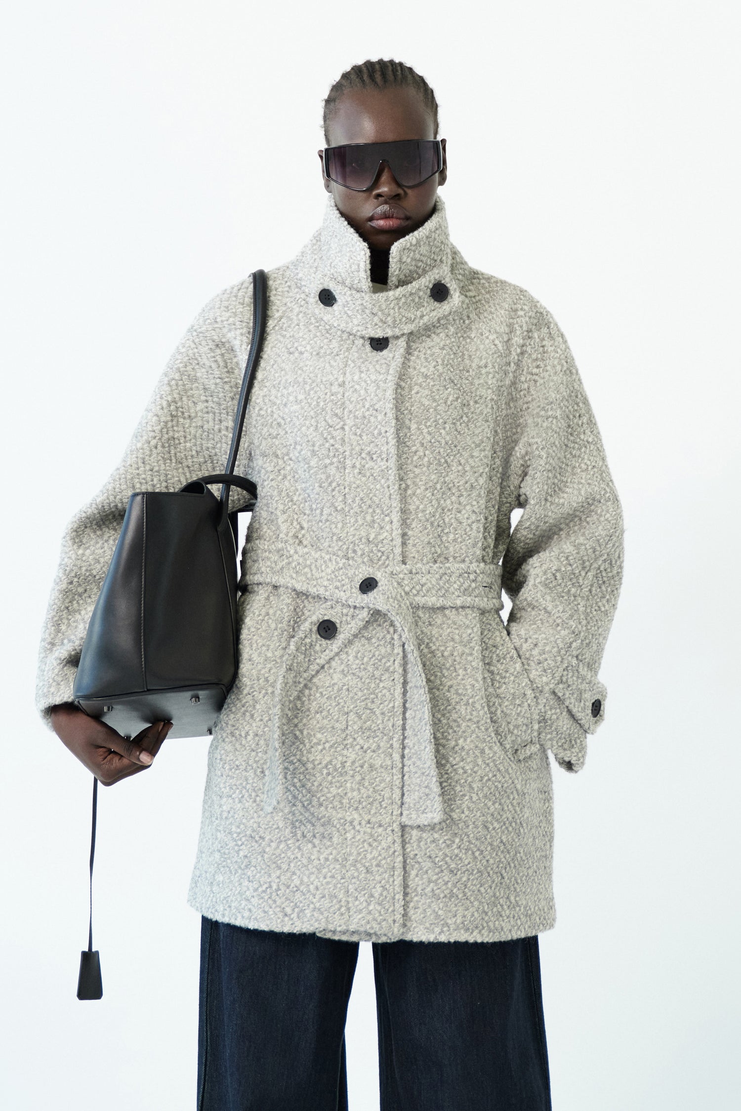 Close-up of the ELEH - THE AURELIA COAT in light gray wool blend, showing a fabric belt and black cuff button, hanging against a white wall.