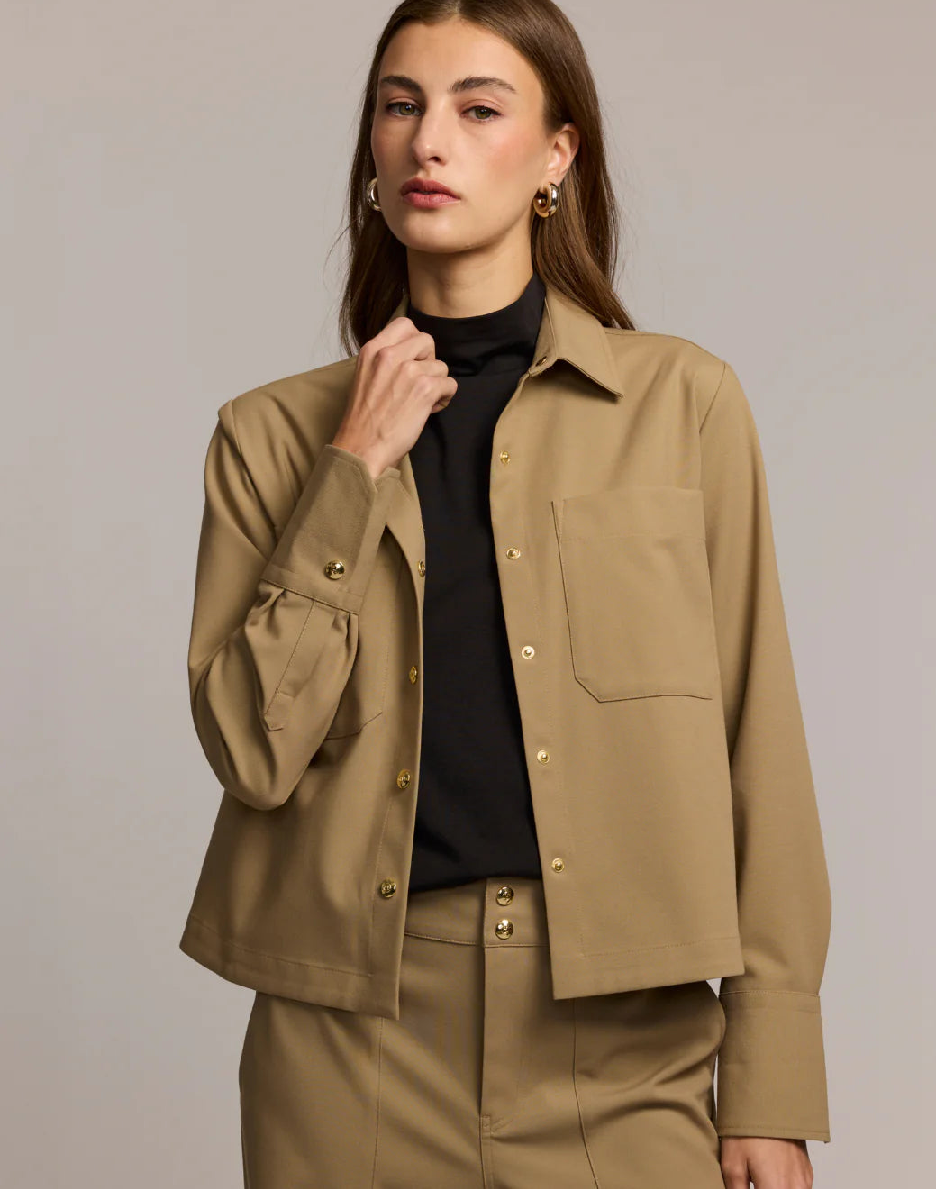 A woman with long brown hair wears a tan button-up jacket over a black top and matching tan pants, standing against a plain beige background. She has a neutral expression and is holding her collar.