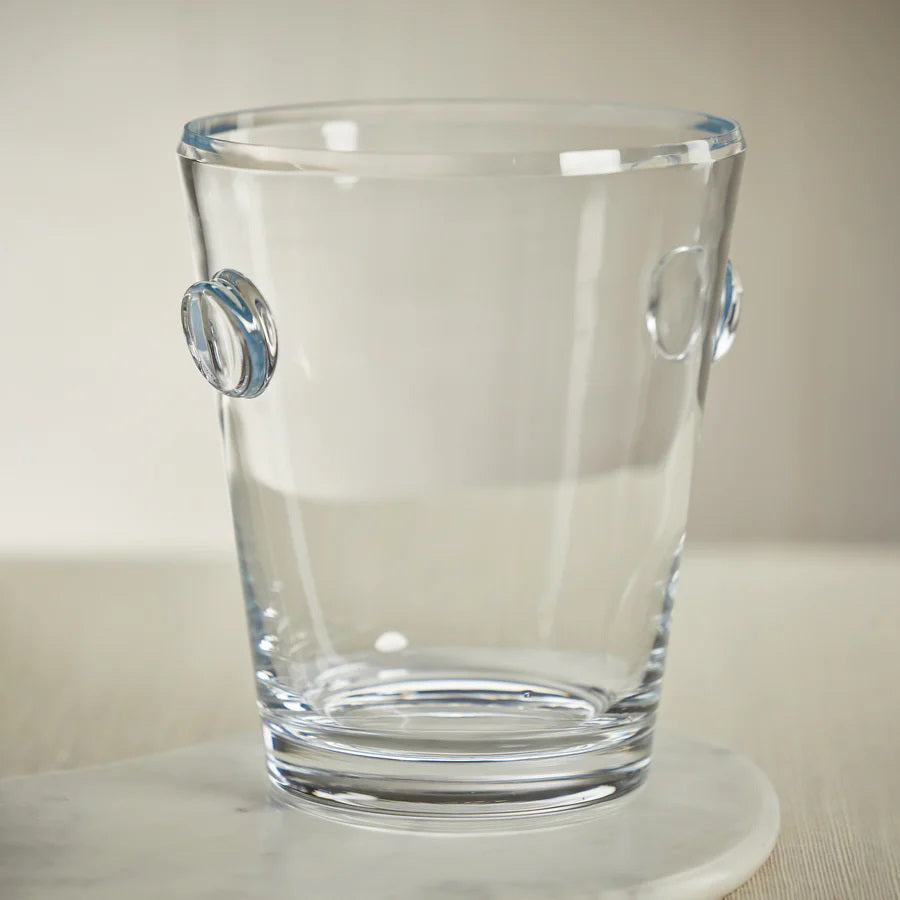 A clear glass ice bucket with two round handles sits on a white marble surface against a neutral background.