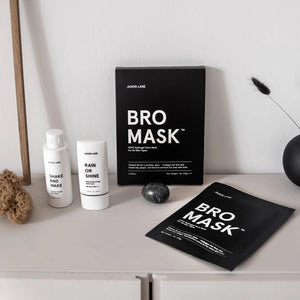 A shirtless man with curly dark hair, a beard, and a chest tattoo lounges outdoors in the sunlight, wearing JAXON LANE's Bro Mask Hydrogel Face Sheet Mask. He holds a drink and relaxes amid greenery, enjoying men's skincare by JAXON LANE.