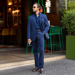 A man in COLLARS AND CO Maverick Performance Pants in Navy, a dark shirt, and sunglasses stands confidently outside a restaurant with green chairs and lush plants nearby, holding his phone.