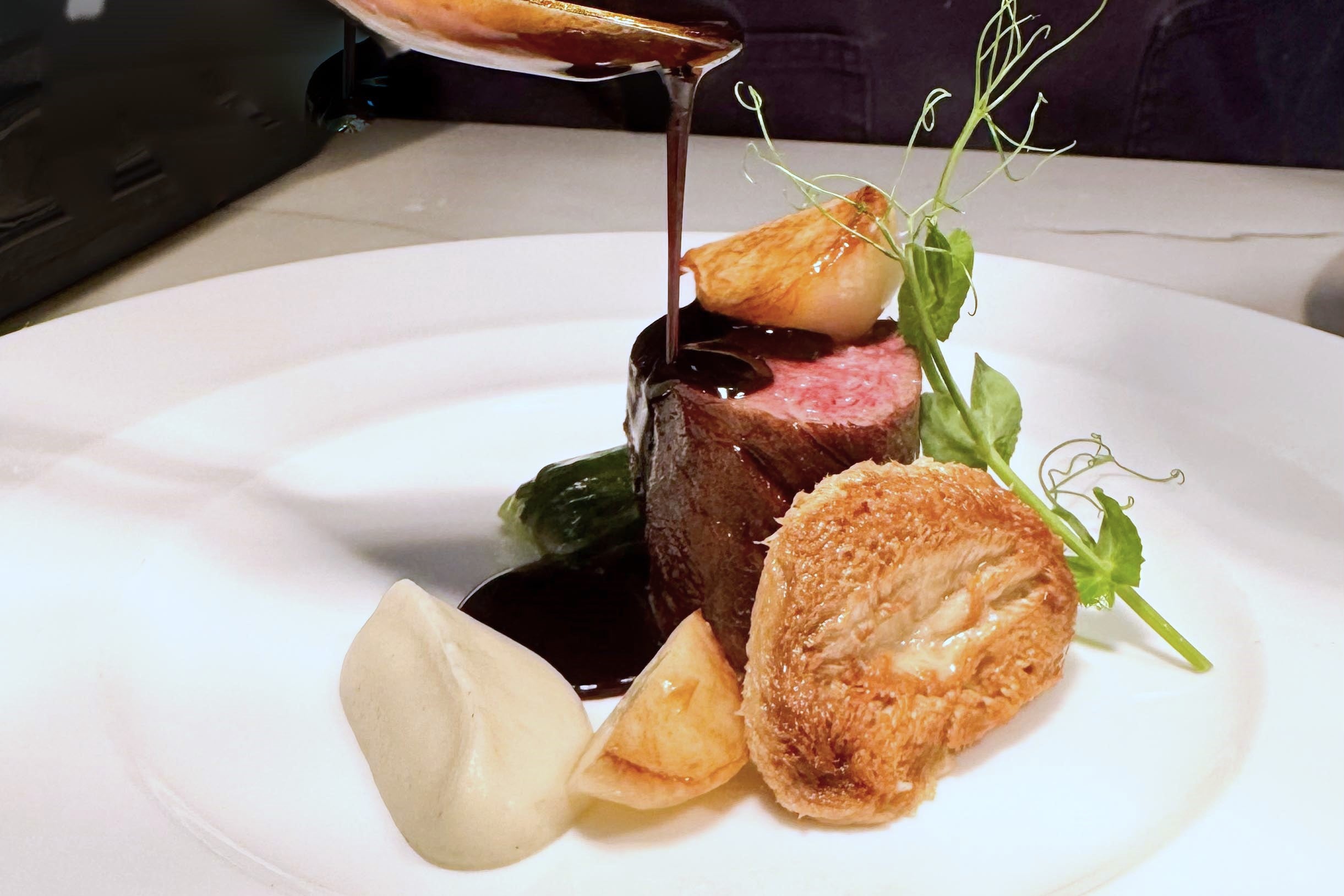 A gourmet dish featuring a medium-rare meat medallion topped with rich brown sauce, surrounded by pureed sauce, fried root vegetables, a green garnish, and a crispy roll on a white plate.