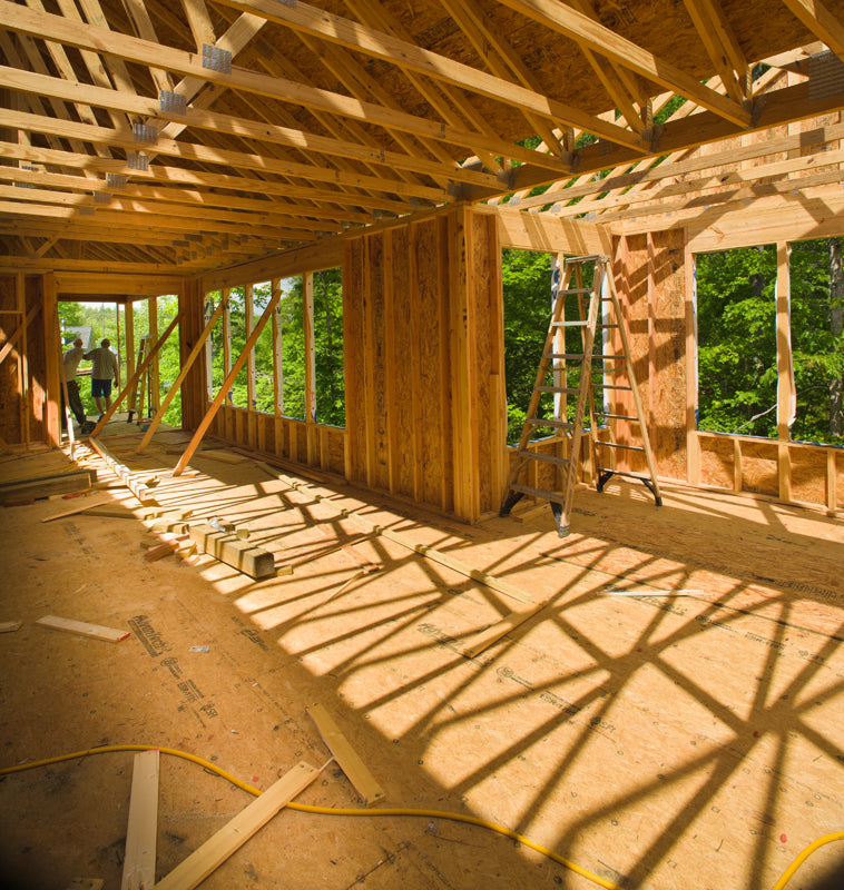 A partially constructed wooden house with exposed framing, open walls, and a ladder inside. Sunlight casts shadows across the floor. Two people are visible in the background near a window opening.