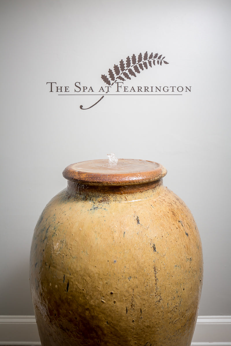 A large rustic water jar fountain sits below a wall sign reading "The Spa at Fearrington" with a fern graphic above the text.