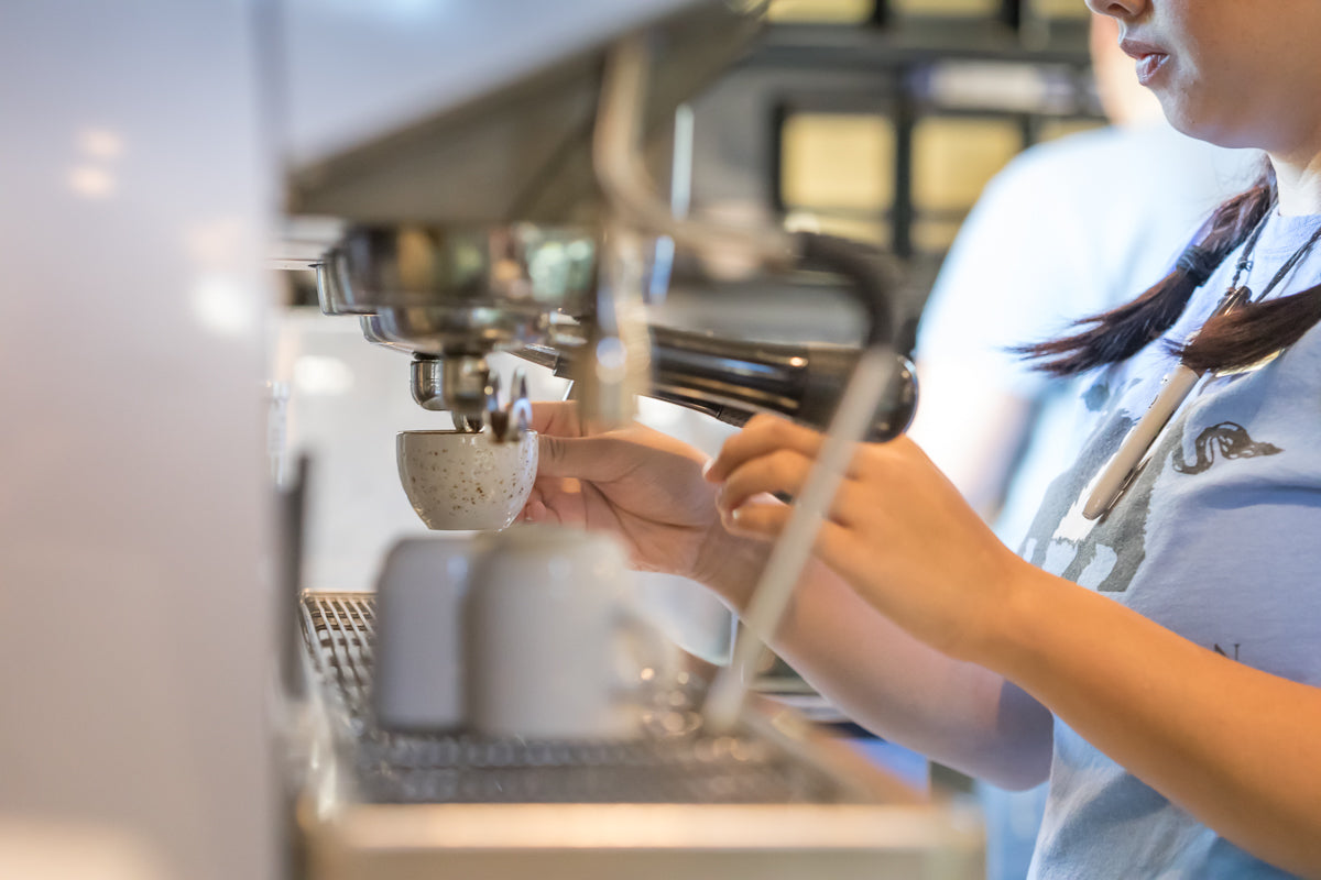A barista is using an espresso machine to prepare coffee, holding a speckled cup under the portafilter. The scene is bright, and the barista wears a light blue shirt.
