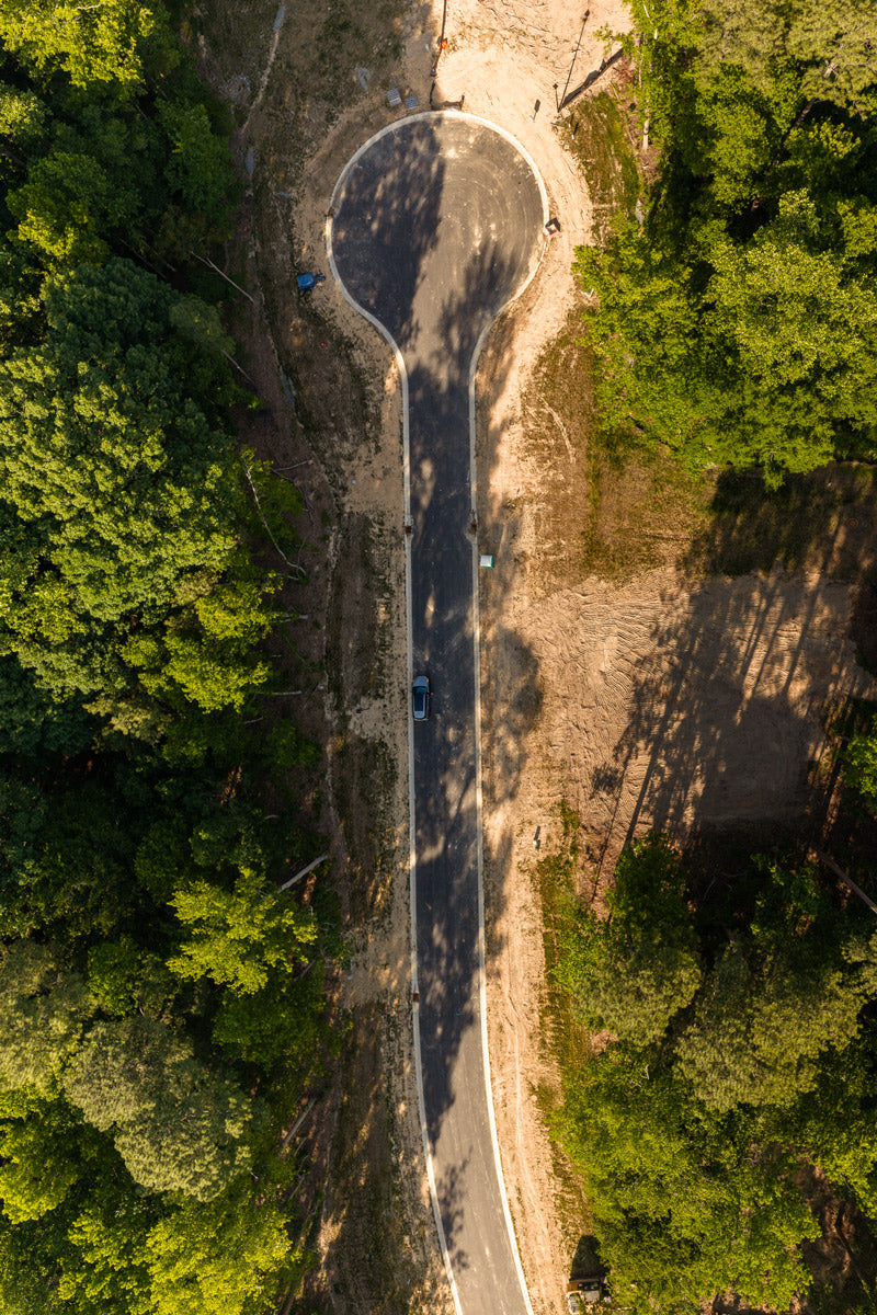 Aerial view of a cul-de-sac surrounded by dense green trees, with a single parked car at the end of the paved road. Sunlight and shadows highlight the forested and cleared areas.