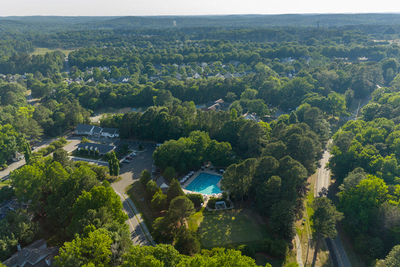 Aerial view of a tree-covered suburban neighborhood with a large rectangular swimming pool in the center, surrounded by greenery, houses, and roads on a sunny day.