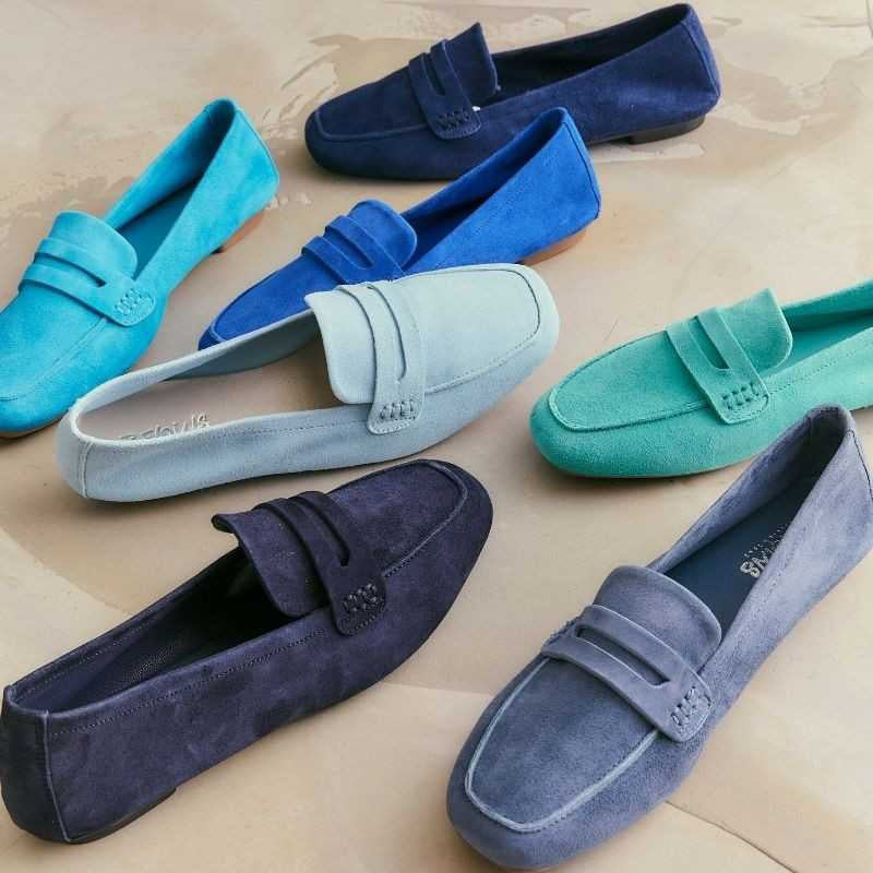A variety of suede loafers in different shades of blue and green are arranged on a light beige surface. The shoes are neatly placed, showcasing their colors and classic design.