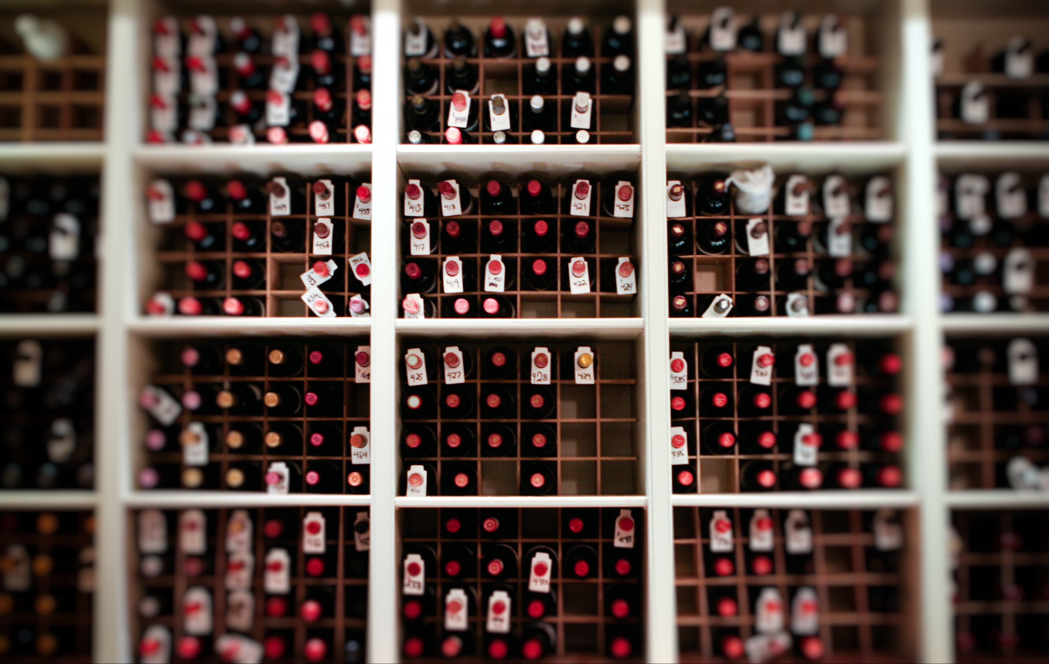 A wall of wine bottles stored horizontally in a grid-like wooden wine rack, each bottle topped with a red or white cap and labeled with hanging tags. The image is slightly blurred at the edges.