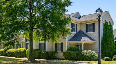 A two-story beige house with dark shutters and a gabled roof is surrounded by green trees and bushes. A black streetlamp stands near the sidewalk under a clear blue sky.