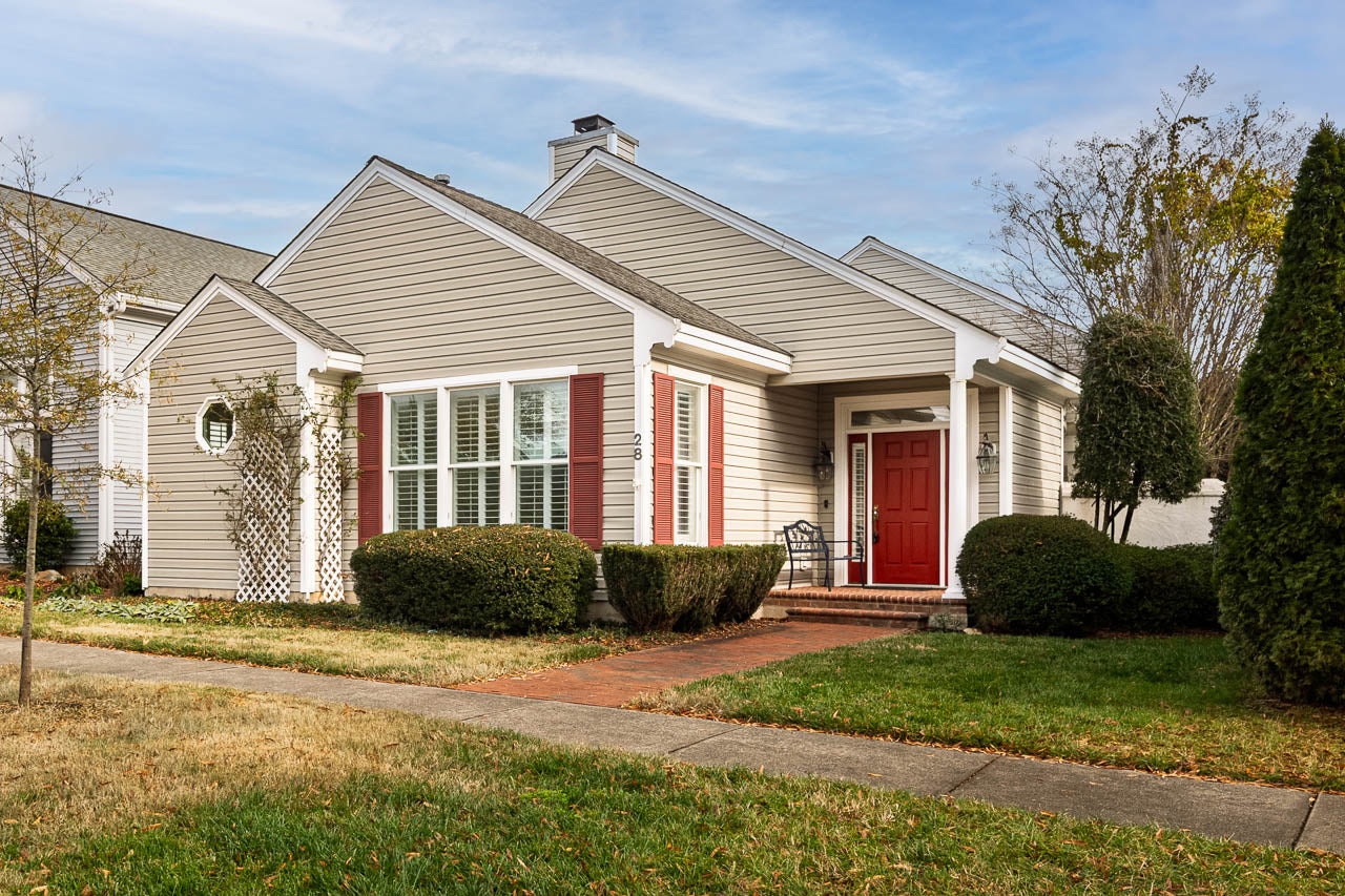 Single-story beige house with white trim, red shutters, and a red front door. Brick walkway leads to the entrance, surrounded by neatly trimmed bushes and lawn, under a partly cloudy sky.