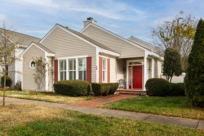Single-story beige house with white trim, red shutters, and a red front door. Brick walkway leads to the entrance, surrounded by neatly trimmed bushes and lawn, under a partly cloudy sky.