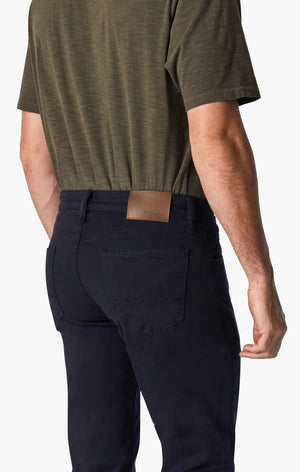 A man with short hair and a beard stands facing forward, wearing an olive green T-shirt, 34 HERITAGE - COURAGE STRAIGHT LEG PANT IN NAVY TWILL, and brown shoes against a plain white background.