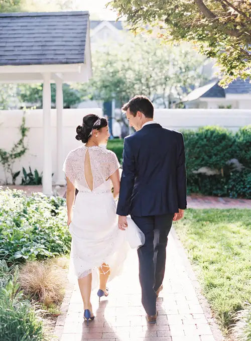 A bride and groom walk hand in hand along a garden path, dressed in wedding attire, with sunlight filtering through trees and greenery surrounding them.