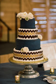 A three-tiered cake with dark blue fondant, gold and white zigzag patterns on each tier, and decorated with white flowers, displayed on a silver cake stand.