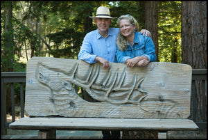 A man in a straw hat and a woman in a denim jacket smile together outdoors by a wooden bench with a stag carving—an inviting scene evoking the Peter Michael Wine Dinner by FEARRINGTON VILLAGE, surrounded by tall trees and greenery.