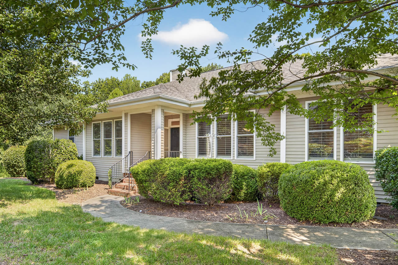 Single-story beige house with white trim, large front windows, and a covered porch. The yard features well-maintained bushes, green grass, and a sidewalk, with tree branches partially framing the scene under a blue sky.