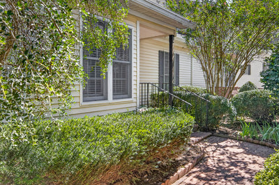 A brick walkway leads to the side entrance of a house with tan siding, large windows, and manicured shrubs.