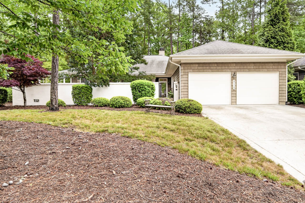 Single-story house with a two-car garage, brown siding, and a manicured yard surrounded by trees and shrubs. Concrete driveway and walkway lead to the front door.