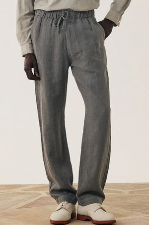A person wearing HARTFORD - LINEN JOGGY PANTS IN ARMY with hands in pockets, a light shirt, and white dress shoes with brown soles stands on a patterned floor.