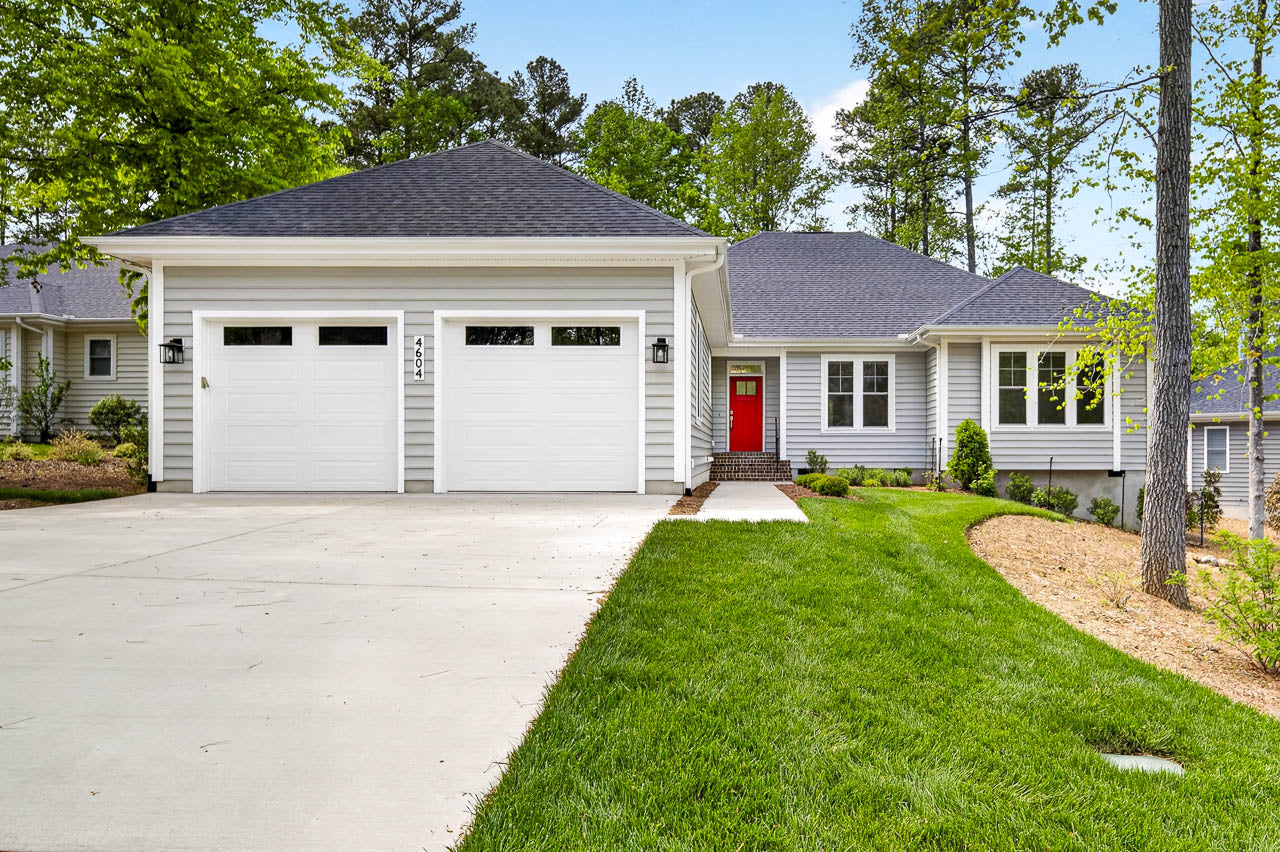 A modern single-story house with gray siding, a dark gray roof, two white garage doors, a bright red front door, and a well-maintained front lawn bordered by trees and shrubs.