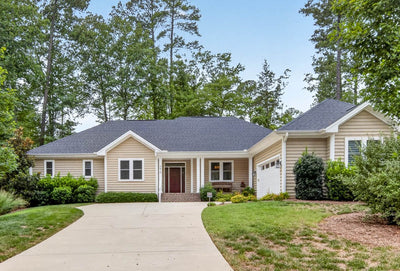 Single-story beige house with a dark gray roof, white trim, two-car garage, and a wide driveway, surrounded by green lawns, shrubs, and tall trees in the background.
