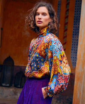 The MOMONI - THIERRY BLOUSE IN PRINTED STRETCH SATIN is a long-sleeved, oversize silk blouse with a multicolored paisley and floral print, high pleated collar, and buttoned cuffs, shown on a plain white background.