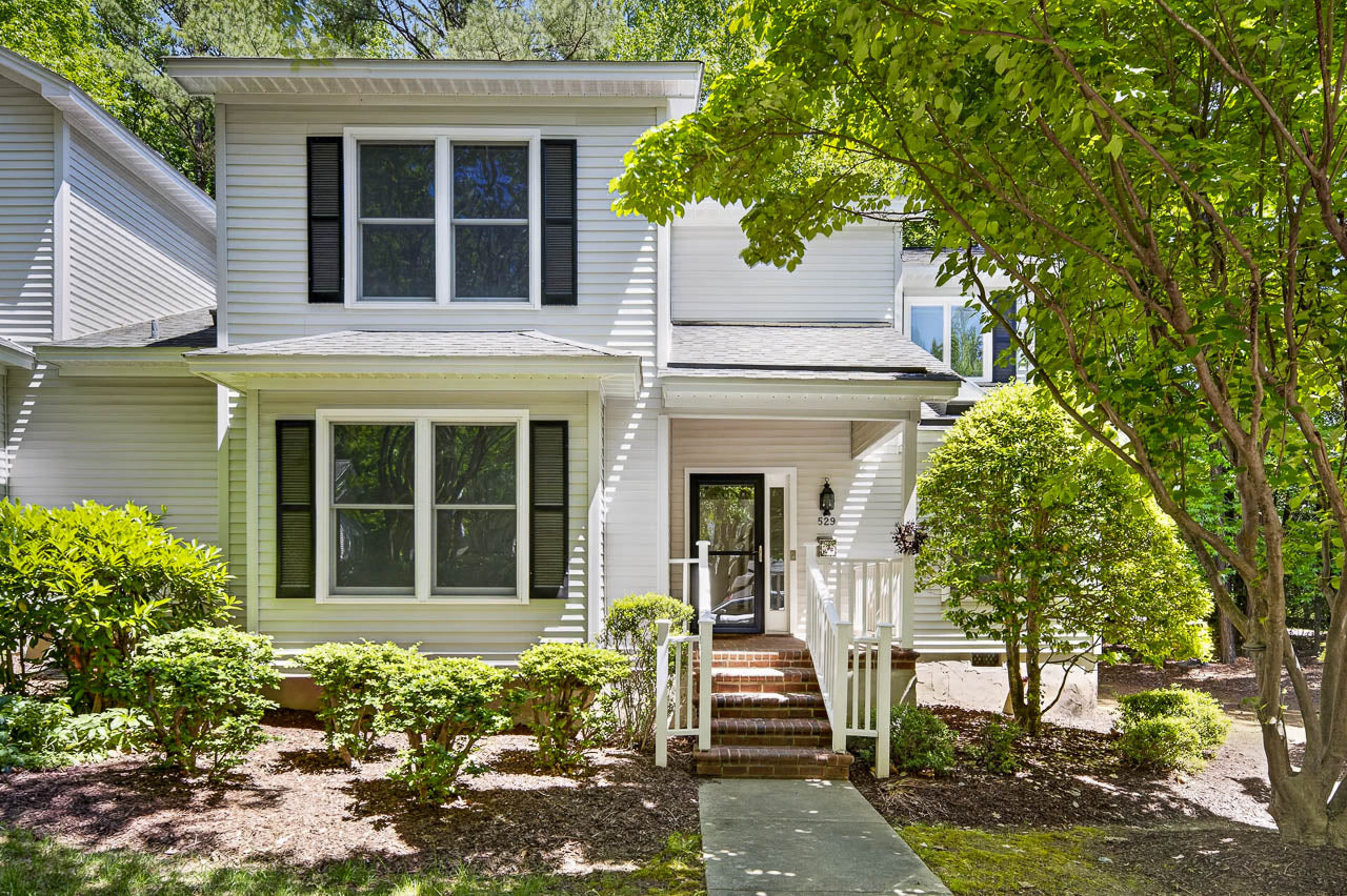 Two-story white house with black shutters, a covered front porch, and brick steps, surrounded by green bushes and trees on a sunny day.